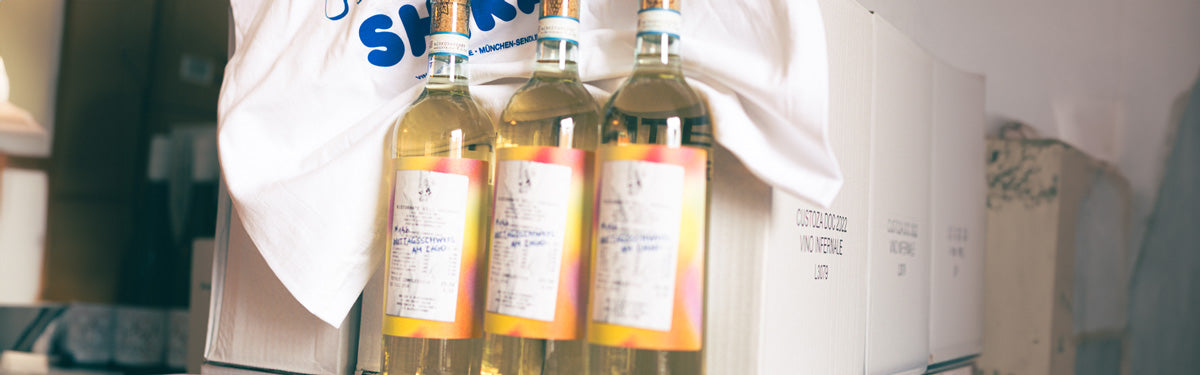 Drei Flaschen Weißwein mit farbigen Etiketten vor weißen Kartons.