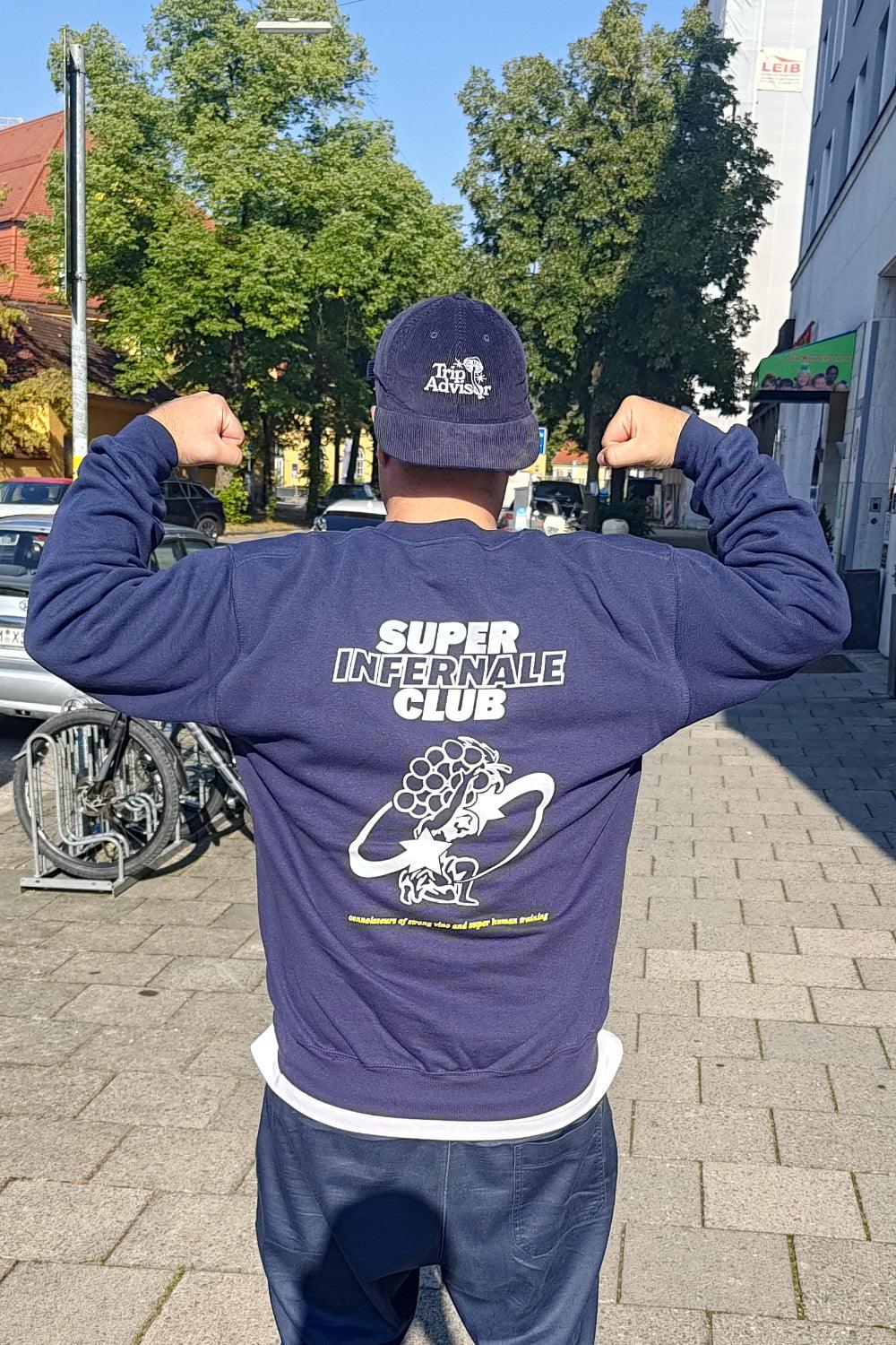 Person mit blauem Super Infernale Club Sweatshirt und Cap zeigt Muskeln im Freien.